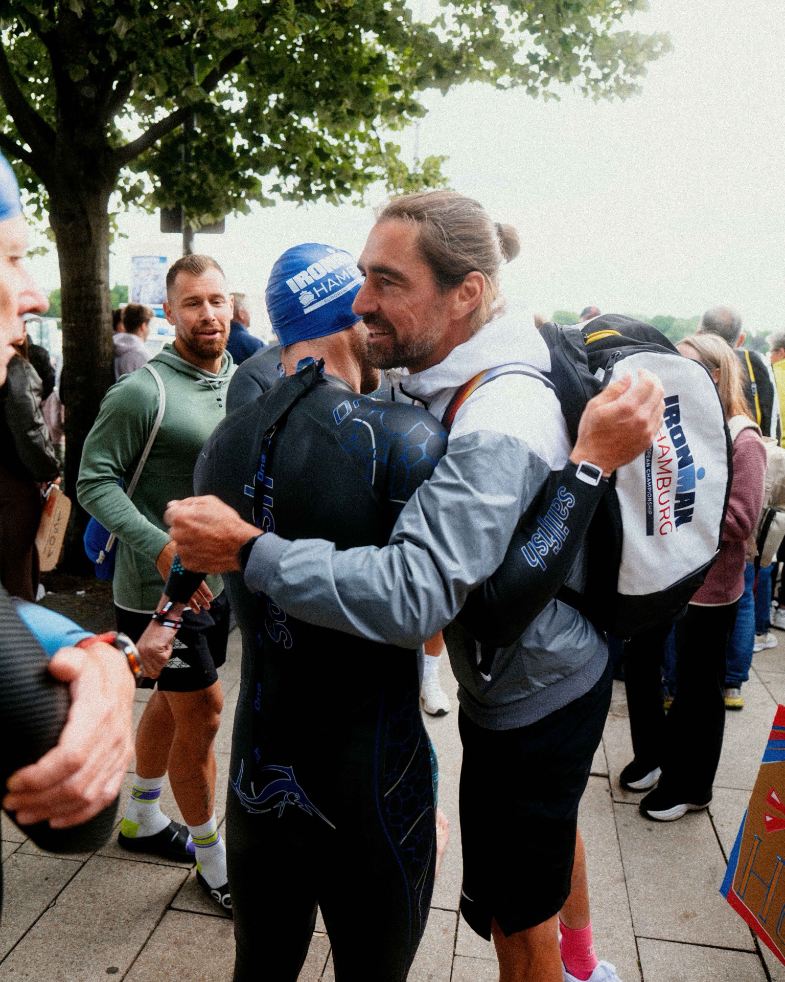 Coach Manuel umarmt einen Athleten im Neoprenanzug beim IRONMAN Hamburg, im Hintergrund Coach Laurenz